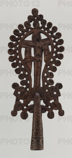 Processional cross, Ethiopian workshop. The pear-shaped frame depicts Christ on the cross with John and Mary standing at the foot of the cross in flat relief. The figures are schematically rendered. Christ is bearded, long-haired, with open eyes; his head is slightly tilted to his right left shoulder. He is dressed only in a loincloth and his feet are nailed with a single nail. The pear-shaped frame is decorated with a braided-rope pattern and bordered along the outer edge with an openwork braided ornament. The cone-shaped sleeve, flattened on the sides, is decorated with a vertical strip of braid and at the bottom with three horizontal strips of checkered, zigzag, and braided pattern. The arms connecting the sleeve with the cross take the form of an openwork braid. 20th century, Ethiopia. Cross; height 40.5 cm, width 16.0 cm., A carved wooden object in dark brown with a tall central figure flanked by two smaller figures, all carved in relief within an oval frame; the frame and surroun...