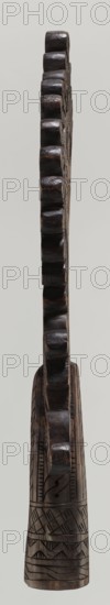 Processional cross, Ethiopian workshop. Pear-shaped frame depicts Christ on the cross with John and Mary standing at the foot in flat relief. Figures are schematically rendered; Christ is bearded, long-haired, with open eyes, head slightly tilted to his right left shoulder, dressed only in a loincloth, feet nailed with a single nail. Pear-shaped frame decorated with braided-rope pattern and bordered with an openwork braided ornament. Cone-shaped sleeve, flattened on the sides, decorated with a vertical strip of braid and at the bottom with three horizontal strips of checkered, zigzag, and braided patterns. Arms connecting the sleeve with the cross form an openwork braid. 20th century, Ethiopia. Cross; height 40.5 cm, width 16.0 cm., A tall, narrow carved wooden object shown in profile with a scalloped edge running vertically; the wood appears dark brown with lighter brown highlights and a slightly glossy surface; the lower portion features carved geometric patterns and linear incisions...