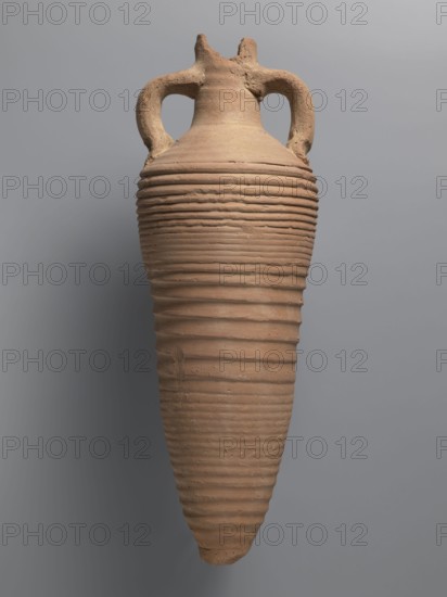 Amphora, unknown Egyptian workshop. Amphora with a spindle-shaped belly, fluted walls, and a pointed tip. The neck is narrow with a reflected rim, elongated, and flows into sloping shoulders. Two cylindrical handles are attached to the neck and shoulders. A hole for wine fermentation is drilled in the neck. The interior is impregnated with resin. Outer surface light brown and smooth; belly ribbed. Made of light brown alluvial Nile clay Naqlun AC1C ceramic fabric, 5th–7th century, Naqlun. Vessel amphora, height 56.6+ cm preserved, neck diameter 6.5 cm, body diameter 20.2 cm., A tall, elongated ceramic vessel with a tapered, pointed base and a wider rounded shoulder, featuring a pair of curved handles near the top; the surface has evenly spaced horizontal ridges encircling the body and a rough, textured finish with some irregularities; the colors visible are terracotta brown on the vessel and a neutral gray background.