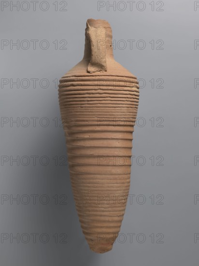 Amphora, unknown Egyptian workshop; amphorae; Egyptian ceramics; storage vessel; Coptic vessel;, amphora; Nile clay. Amphora with a spindle-shaped belly, fluted walls, and a pointed tip. Narrow neck with reflected rim, elongated and flowing into sloping shoulders. Two cylindrical vices attached to the neck and shoulders. A hole for wine fermentation drilled in the neck. Interior impregnated with resin. Outer surface light brown and smooth; ribbed belly. Made of light brown alluvial clay Naqlun AC1C ceramic fabric, 5th–7th century, Naqlun findspot. Vessel amphora, height 56.6+ cm preserved, neck diameter 6.5 cm, body diameter 20.2 cm., A tall, elongated ceramic vessel with a tapered base and a narrow top, showing a rough, textured surface with horizontal ridges running along its body; the vessel is primarily terracotta in color with variations of light brown and beige, and it rests against a smooth gray background.
