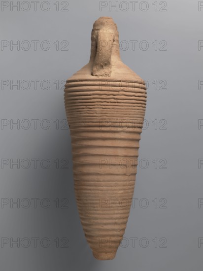 Amphora, unknown Egyptian workshop. Amphora with a spindle-shaped belly, fluted walls, and a pointed tip. Narrow neck with reflected rim, elongated and flowing into sloping shoulders. Two cylindrical handles attached to the neck and shoulders. A hole for wine fermentation drilled in the neck. Interior impregnated with resin. Outer surface light brown and smooth; belly ribbed. Made of light brown alluvial Nile clay Naqlun AC1C ceramic mass, 5th–7th century, Naqlun. Vessel amphora, preserved height 56.6+ cm; neck diameter 6.5 cm; body diameter 20.2 cm., A tall, elongated clay vessel with a narrow, rounded base and a wider shoulder, displaying horizontal ridged bands that wrap around its body; the surface is textured with visible irregularities and a rough patch at the top where a short handle or neck extends upward; the vessel appears in shades of terracotta and light brown against a smooth grey background, with subtle shadowing creating darker grey and muted brown tones along one side.