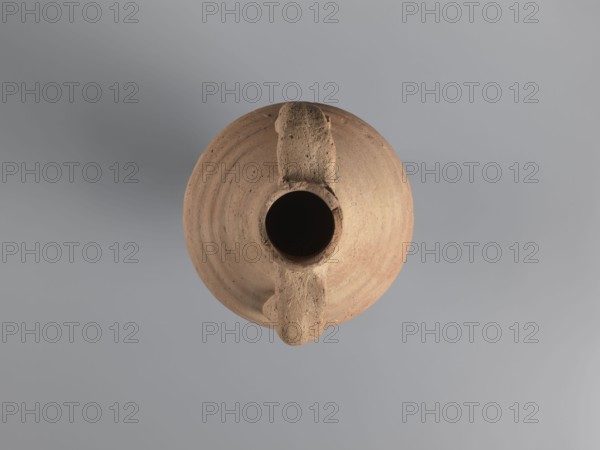 Amphora, unknown Egyptian workshop. Amphora with a spindle-shaped belly, fluted walls, and a pointed tip. Narrow neck with a reflected rim, elongated and flowing into sloping shoulders. Two cylindrical handles attached to the neck and shoulders. A hole for wine fermentation drilled in the neck. Interior impregnated with resin. Outer surface light brown and smooth; ribbed belly. Made of light brown alluvial Nile clay Naqlun AC1C ceramic fabric. Function: storage vessel Coptic amphora. Dated 5th–7th century. Findspot: Naqlun. Height 56.6+ cm preserved; neck diameter 6.5 cm; body diameter 20.2 cm., A top-down view of a round clay vessel with a central circular opening and a pronounced handle-like ridge running across its diameter; the vessel shows concentric rings and a textured surface with small pits and irregularities. The colors visible are shades of brown on the vessel and shades of gray in the background.