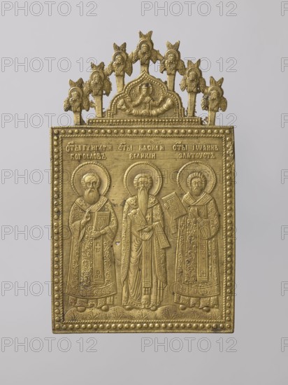 Icon: Holy Fathers of the Church. Old Believers workshop. Metal icon showing Saints Gregory the Theologian, Basil the Great, and John Chrysostom in full-figure, frontal views. They are dressed in bishops' robes, holding books, standing on flower-covered ground. Identifying inscriptions above them. Background horizontally engraved with thin lines. A ribbon ornament with raised dots surrounds the composition. On the icon's axis is a medallion topped with a kokoshnik-shaped arch depicting Christ as the High Priest, blessing with both hands. Cherubim and ripidions form a semicircular arch around it. Relief is precise and meticulous, with ornamentation and careful detail. 19th century, Russia. Materials: metal. Dimensions: height 19.8 cm, width 12 cm, depth 0.2 cm., A rectangular metal plaque with a decorative arched top, rendered in a gold color, showing three standing robed figures with halos, each holding a book or object, framed by a beaded border and a patterned lower edge; above the f...