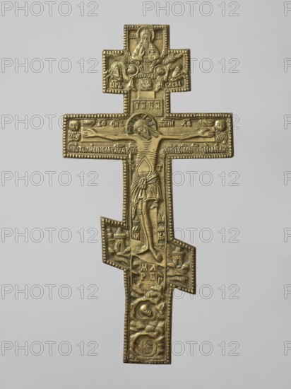 Cross: Crucifixion of Christ, Old Believer Popovtsy workshop; eight-pointed cross with deep relief, schematic, somewhat archaizing style. On the cross: image of the Crucifixion; below, a grotto with Adam's skull and the tree of Seth; at the footstool level, a view of Jerusalem; on either side of Christ on a long beam, the sun and the moon; in the upper part, a bust-like image of the Lord of Hosts with the dove of the Holy Spirit below. Reverse engraved with a prayer to the Holy Cross and an image of an eight-pointed cross. Iconography includes Lord of Hosts, Jesus Christ, God the Father, the moon, Crucifixion, the sun, Adam, Golgotha, skull, instruments of the Passion, Jerusalem, the Holy Spirit, ornaments, inscriptions, halos. 19th century, Russia. Dimensions: height 34.3 cm, width 17.1 cm., A metal cross-shaped object in metallic gold tones with darker gold and brownish shadows, featuring a central raised figure with arms outstretched, surrounded by a few smaller raised figures and s...
