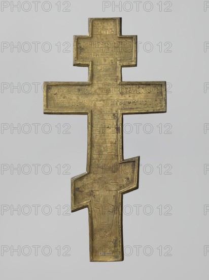 Cross: Crucifixion of Christ. Old Believers workshop. Brass cast eight-pointed cross with rough casting; background enamel in navy blue, sky blue, and white. Obverse: image of the Crucifixion — Christ suspended on the cross; below, a grotto with Adam's skull and the tree of Seth; at the footstool level, a view of Jerusalem; on the long beam beside Christ, the sun and the moon; in the upper part, a bust-like image of the Lord of Hosts with the dove of the Holy Spirit below; ornaments, inscriptions, and halo. Reverse: engraved prayer to the Holy Cross and an image of an eight-pointed cross. Iconography: Jesus Christ; Lord of Hosts; God the Father; Holy Spirit; Crucifixion; Adam; Golgotha; skull; instruments of the Passion; sun; moon; Jerusalem; ornaments; inscriptions. 19th century, Russia. Dimensions: height 34.3 cm, width 17.3 cm., A metal cross with an elongated shape and a stepped, off-center lower arm, shown against a plain light gray background; the cross has a weathered golden-bro...