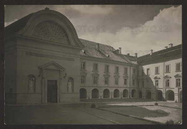 Vilnius. Stefan Batory University - Skarga Courtyard, Bulhak, Jan 1876-1950 photographer, Vilnius Lithuania, ca. 1934, photography, nature photography, height 11.3 cm, width 16.7 cm, A sepia-toned courtyard scene shows a large classical building with arched openings along the ground level and rectangular windows on the upper floor, a curved pediment with a fan-shaped window above a decorated entrance, and two niches flanking the doorway each containing a standing statue; a paved drive and formal lawn with trimmed hedges and planting beds occupy the foreground, and the building's sloped roof with small chimneys and dormers is visible under a sky with scattered clouds.
