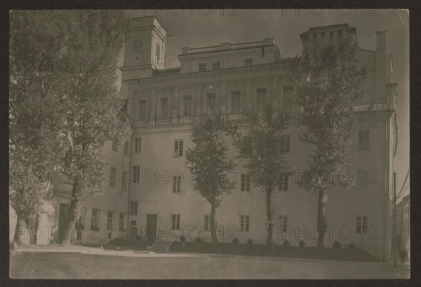 Vilnius. Stefan Batory University — From Napoleon Square. Photographer: Jan Bulhak 1876–1950. Vilnius Lithuania; ca. 1934. Photography; nature photography. Image dimensions: height 11.3 cm, width 16.7 cm., A large multi-story building with a tall tower at one corner stands behind a row of several tall trees; the building has many windows and decorative architectural details along its upper facade, and there are a few doorways and small steps at ground level. The scene shows a paved area or street in front of the building and a narrow strip of landscaping with low shrubs near the base of the building. The colors visible are shades of sepia, brown, beige, and muted gray-green.