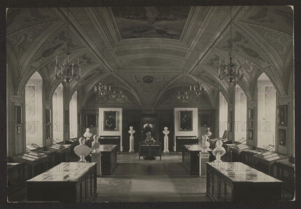 Vilnius. Stefan Batory University - Library Room, Bulhak, Jan 1876-1950 photographer, interiors, library interiors, ca. 1934, Vilnius, photography, nature photography, height 11.4 cm, width 16.8 cm, A large ornate interior room with a vaulted ceiling featuring decorative plasterwork and painted panels, several hanging chandeliers, tall arched windows along both side walls, rows of display cabinets with glass tops running down the length of the room, and a group of sculpted busts mounted on pedestals positioned on and beside the display tables; framed portraits hang on the far wall between decorative panels, and the visible colors are shades of gray, black, white, and sepia tones.