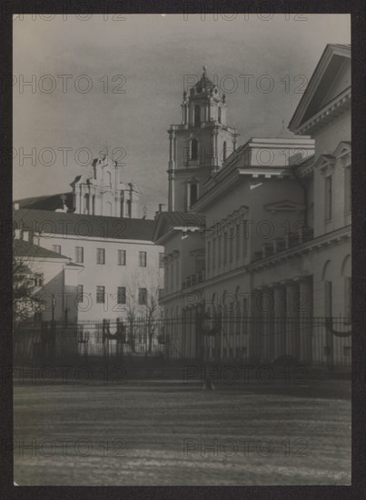 Vilnius. Bishop's Palace later Representative Palace and Church of St. John the Baptist and St. John the Evangelist. Photograph by Jan Bulhak 1876–1950, ca. 1937. Photography; nature photography. Height 15.7 cm, width 16.8 cm., A grayscale photograph showing a row of classical buildings with columns and decorative rooflines, a taller ornate tower rising behind them, and a separate ornate façade further in the background; a dark iron fence with decorative elements runs across the foreground, and leafless trees stand behind the fence; the sky appears clear and pale, and the image contains shades of black, gray, and white.