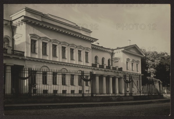 Vilnius. Bishop's Palace from Napoleon Square, photographed by Jan Bulhak 1876–1950, Vilnius Lithuania; architecture: Bishop's Palace later Representative Palace, Vilnius, ca. 1937; photographer: Mieczyslaw Orlowicz 1881–1959; photography, nature photography; height 11.3 cm, width 16.8 cm, A large classical building with a long facade and decorative cornices is shown behind a tall metal fence; the building is light in color with darker window frames and trim, featuring a row of rectangular windows with pediments on the upper level and arched windows and columns on a lower section, and a portico with columns on the right side. The fence in front is dark and ornate with vertical bars and decorative swags, and there is a cobblestone street in the foreground. Trees with dark foliage are visible behind and to the right of the building, and the sky above is light and mostly clear. The overall image tones are shades of black, white, and gray, with contrasts between the light facade, dark fenc...