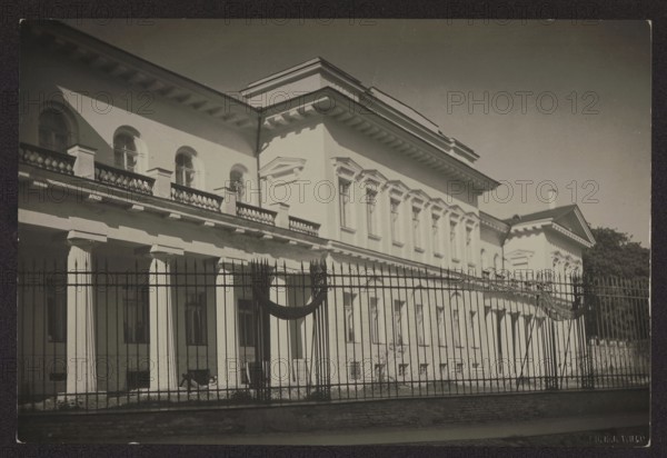 Vilnius. Bishop's Palace from Napoleon Square, photograph by Jan Bulhak 1876–1950. Bishop's Palace later Representative Palace, Vilnius, ca. 1934. Photography; nature photography. Height 11.3 cm, width 16.6 cm., A large classical building with tall columns and decorative cornices is shown behind a tall metal fence; the façade appears light in color with shadows creating contrast, and the roofline and window frames are visible, while the fence in front is dark and vertical with pointed tops and a draped dark fabric element; the image shows areas of light and shadow, with tones of black, white, gray, and variations of sepia across the photograph, and some trees or foliage are visible at the far right.