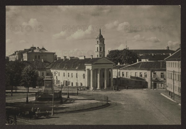 Vilnius. Napoleon Square. Photographer: Jan Bulhak 1876–1950. Orlowicz, Mieczyslaw 1881–1959 - architecture. Napoleon Square Vilnius, city square; photograph taken in 1915; in the middle of the square the pedestal of the Muravyev monument, ca. 1934. Place: Vilnius Lithuania. Photograph; nature photograph. Height 11.0 cm, width 16.2 cm., The image shows an outdoor urban scene in muted grayscale tones with a group of classical buildings, including one with prominent columns and a triangular pediment, a tall bell tower with a spire rising behind it, and several low-roofed structures arranged around an open paved square. In the foreground there is a fenced monument base on a small raised grassy area with a few trees nearby, street lamps, and a bench with a few people seated. The sky above contains scattered clouds, and the overall colors visible are shades of black, white, and gray.