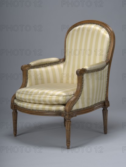 Armchair, covered with striped fabric with a loose cushion; wooden parts waxed. 4th quarter of the 18th century. Armchair, height 93.5 cm, width 73.4 cm, depth 62 cm., An upholstered armchair with a wooden frame showing a carved curved back, arms, and tapered legs, covered in striped fabric in shades of cream and pale yellow with brass-colored nailhead trim along the edges; the wood appears medium brown and the background and floor are shades of gray.
