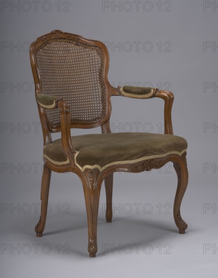 Armchair, In the upper part of the frame a motif of interwoven flowers; upholstered seat; 2nd half of the 18th century; Poland; armchair; height 88 cm, width 63 cm, depth 48 cm., The image shows a wooden armchair with a carved frame and a woven cane back, featuring a curved top rail and scrolling arms; the seat and small padded armrests are upholstered in an olive green fabric with a light trim, and the wood has a medium brown finish with carved decorative details on the apron and legs; the background is a smooth gray.