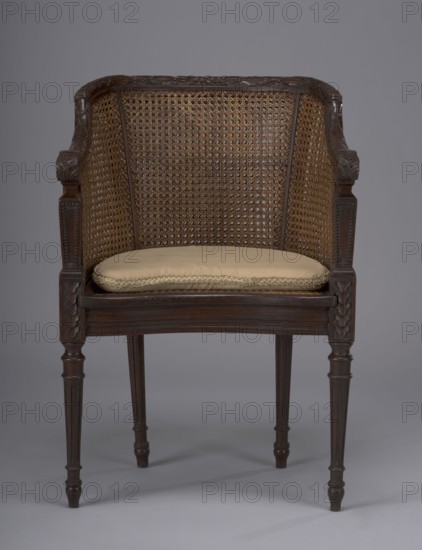 Armchair in the Louis XVI style; seat and backrest woven; 18th/19th century; height 81 cm, width 55 cm, depth 50 cm;, A wooden chair with a woven cane back and sides and carved wooden arms and legs, featuring a light beige cushion on the seat; visible colors include brown tones in the wood and cane, beige on the cushion, and a gray background.