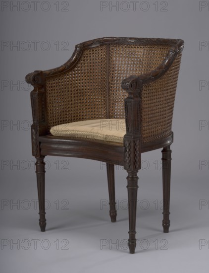 Armchair, Louis XVI style, seat and backrests woven, 18th/19th century; height 81 cm, width 55 cm, depth 50 cm., A wooden chair with a curved cane back and sides, featuring carved arm supports and decorative carved details on the frame, a light beige cushion on the seat, and tapered turned legs; visible colors include brown in various tones on the wood and cane, beige on the cushion, and a neutral gray background.