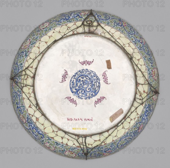 Platter, Large decorative platter with flat bottom and edges rising in an arc, set on a low, wide ring foot. Obverse: figural composition in a large tondo overlapping the lower parts of the slightly rising edges — a genre scene of scholars taking a siesta on a sunny summer day on a terrace of a landscape park. High foundations of decoratively bricked boulders rise above the valley of a large river. The terrace with an openwork balustrade is overgrown with grass, large deciduous trees, and banana trees nestled among rocks and boulders. In the center of the terrace is a large table with three scholars seated and a girl beside them. Reverse: mirrored leg decoration — a decorative composition of five pink bats surrounding a blue dragon, combined with plant motifs. 19th century, China. Plate; height 5.8 cm, diameter 44 cm., A round ceramic plate viewed from above with a central circular medallion decorated in blue surrounded by a group of ornamental pink motifs on a white background; the ri...