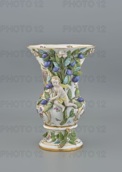 Vase, 19th/20th century, Meissen, vase, diameter 36 cm, diameter 24 cm base, The image shows a decorative porcelain vase with a flared rim and a rounded body on a pedestal, predominantly white with accents of gold, green, blue, purple, yellow, and pink; the vase is embellished with raised green leaves and clusters of blue-purple fruit, small white flowers, and painted floral motifs, and features a small sculpted cherub figure in pale flesh tones with yellow cloth elements clinging to the foliage; gold trim highlights the rim and base, and the background is a uniform grey.