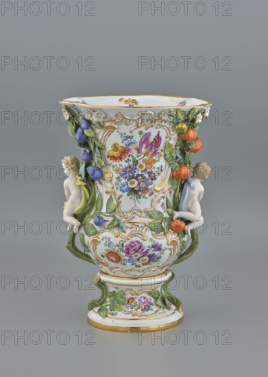 Vase, 19th/20th century, Meissen; vase; diameter 36 cm, base diameter 24 cm., An ornate ceramic vase with a white base decorated with gold accents and colorful painted floral designs, featuring sculpted green leafy garlands and clusters of purple and orange fruit; two small white cherub-like figures cling to the sides, each with subtle skin tones and pale hair, one draped with a pale yellow cloth and the other with a pale blue cloth; the vase has raised gold scrollwork framing panels of multicolored flowers in shades of purple, pink, yellow, orange, blue, and red, and sits on a round white pedestal base with green leaf appliqués and a gold rim, all shown against a plain gray background.