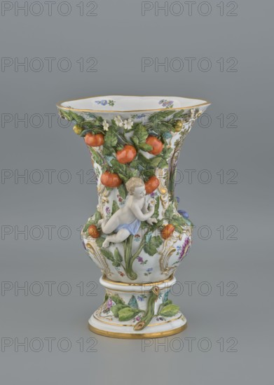 Vase, 19th/20th century, Meissen; vase; diameter 36 cm, base diameter 24 cm., A porcelain vase with a flared rim and pedestal base decorated with applied sculptural elements, featuring a reclining cherub figure in pale skin tones with a light blue drapery, surrounded by molded green leaves and clusters of round orange fruit, with small white blossoms and additional floral sprays painted in colors including purple and blue; the vase has gold-painted edging and accents and a white background, photographed against a smooth medium gray backdrop.
