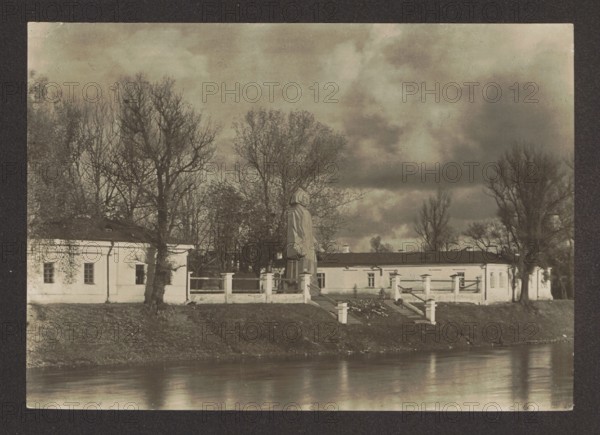 Vilnius. The Neris River and the Mickiewicz Monument, Bulhak, Jan 1876-1950 photographer, monuments, Vilnius Lithuania, Pronaszko, Zbigniew 1885-1958 - reproduction, Mickiewicz, Adam 1798-1855 - iconography, sculpture artist, The Neris River Lithuania; river, rivers, ca. 1934 collected, Vilnius place of creation, photography, nature photography, height 11.5 cm, width 16.3 cm, A sepia-toned photograph showing a riverside scene with a reflective water surface in the foreground and a raised grassy embankment behind it; atop the embankment is a light-colored long building with a low roof and a series of windows, interrupted by a central area with steps and a platform where a tall statue draped in cloth stands; a group of bare and sparsely leaved trees rise behind and around the building, their branches dark against a cloudy sky; the overall palette includes brown, beige, cream, gray, and muted black tones.