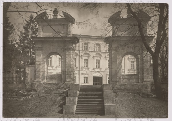 Gate of the Sapieha Palace in Antakalnis, photographed by Jan Bulhak 1876–1950, Vilnius, ca. 1934. Photograph, nature photograph; height 11.6 cm, width 16.5 cm., A sepia-toned photograph shows the front of a large classical building framed by two textured stone gate pillars with arched openings; a flight of stone steps with rectangular block railings leads up between the pillars toward the building. The central façade visible through and beyond the arches displays multiple windows and decorative architectural details. Bare tree branches arch across the top of the image and a few trees flank the scene, with a wooden fence partly visible on one side. The ground in front of the steps appears uneven and covered with dirt and sparse grass. The predominant colors are sepia tones, brown, and shades of beige.