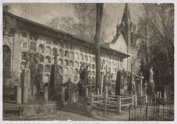 View of the Catacombs, photograph by Jan Bulhak 1876–1950, ca. 1934. Rasos Cemetery Rossa, Vilnius, showing cemetery catacombs. Photograph, 11.3 × 16.4 cm., The image shows an outdoor scene of a cemetery with a long wall of arched niches running across the background and a tall church or chapel with a pointed roof and spires rising behind it. In front of the niche wall are several grave markers and monuments including a few upright stone crosses, a tall dark obelisk, and a variety of headstones and fenced plots. There is a statue of a standing figure near the right side and several wreaths or floral arrangements placed on or near some graves. Trees with bare branches rise above and between the monuments, and low metal and wooden railings form enclosures around some plots. The visible colors are shades of gray, brown, black, and muted beige.
