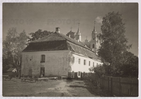 Calvary, Bulhak, Jan 1876–1950 photographer, Vilnius, ca. 1934. Calvary of Vilnius Werki; churches; architecture; sacred architecture; Vilnius Baroque style; photography; nature photography. Image size: height 11.5 cm, width 16.5 cm., A large light-colored two-story building with a steep dark roof fills the foreground, showing a worn exterior and several windows; a small door is visible on the nearer wall. A dirt path and grassy area lead up to the building, bordered by a wooden fence and a few low benches or rails. Tall trees with leafy canopies stand to the left and right of the building, casting shadows. Behind the building rise ornate towers with pointed spires partly obscured by the roof and trees. The scene appears in muted tones of black, white, gray, and sepia.