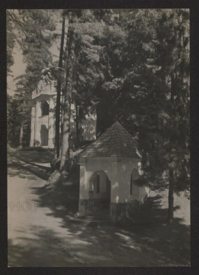 Vilnius vicinity. Calvary — Station. Photographer: Jan Bulhak 1876–1950. Vilnius Calvary Werki. Sacred architecture; Stations of the Cross chapels. Photographer Mieczyslaw Orlowicz 1881–1959 involved. Circa 1934. Photography, nature photography. Dimensions: height 16.3 cm, width 11.6 cm., A grayscale photograph showing a small octagonal pavilion with a shingled pyramidal roof and open arched sides set among tall trees, with dappled light and shadow on its pale walls and the ground; a path or driveway curves past the pavilion toward a larger building partly visible through the trees in the background, the larger structure having tall vertical openings and pale facades; the scene contains a group of tall coniferous trees with dense dark foliage, trunks casting long shadows across the ground, and overall tones of black, white, and various grays.