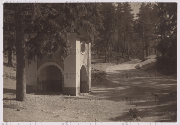 Calvary, Vilnius vicinity; photographer Jan Bulhak 1876–1950; ca. 1920; subject: Calvary of Vilnius Werki, architecture—chapels, village roads, trees; nature photography; dimensions: 11.2 × 16.3 cm., A small building with rounded arch openings and circular windows stands partially shaded beneath tall evergreen trees beside a winding dirt path that curves into a forested area; the scene shows grassy and bare earth foreground with scattered shadows, tree trunks, dense foliage, and distant trees lining the path, with visible colors including shades of brown, tan, cream, and dark green.