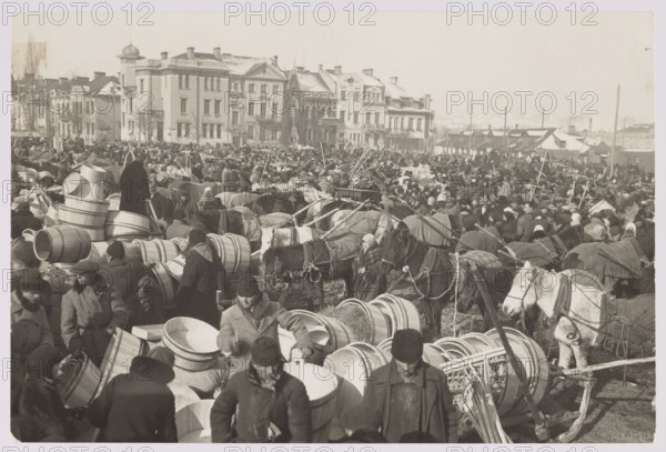 Vilnius. Kaziuk Fair. Photographer: Jan Bulhak 1876–1950. Vilnius Lithuania, Kaziuki fair, photographed circa 1934. Photography; nature photography. Image dimensions: 11.3 cm x 16.8 cm., A crowded outdoor scene filled with a large group of people bundled in heavy coats and hats, surrounded by many wooden barrels and round wooden tubs stacked and scattered in the foreground; several horses and packs appear harnessed and standing among the crowd, some covered with blankets or cloths; a group of multi-story buildings with varied architectural details forms the background skyline, with rooftops and chimneys visible; the overall tones are shades of black, white, and gray creating a monochrome appearance, with textures of wood, fabric, and masonry evident throughout.