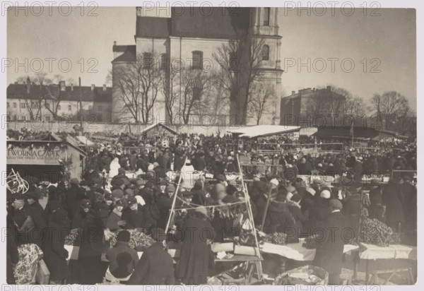 Vilnius. Kaziuk Fair. Photographer: Jan Bulhak 1876–1950. Vilnius Lithuania, Kaziuki fair, photographed ca. 1937. Photography, nature photography. Image dimensions: height 11.2 cm, width 16.5 cm., The image shows a crowded outdoor market scene in muted sepia tones with shades of brown, beige, gray, and black; many people wearing dark coats and hats gather around makeshift wooden stalls and tables covered with cloths, while a few lighter-colored garments and head coverings stand out among the crowd; in the foreground are several tables displaying piles of goods and hanging items on simple frames, and market vendors and shoppers form a dense group extending into the middle ground; behind the market are more stalls with pitched roofs and canopies and a low wall, and in the background a large light-colored stone building with tall windows and towers rises above leafless trees against a pale sky.