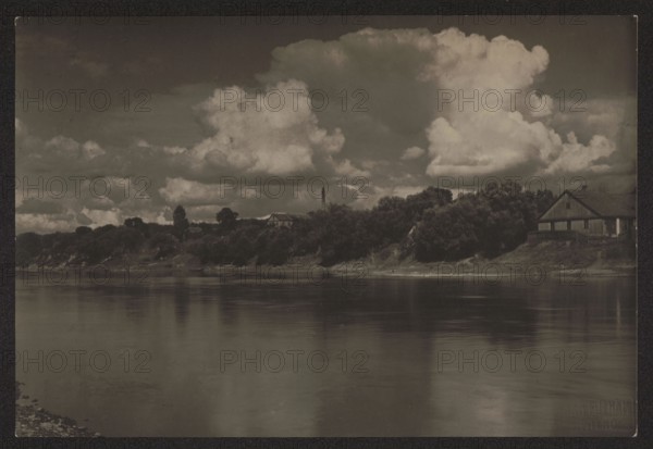 Wilno Vilnius, Wilia in Antakalnis; photographer: Jan Bulhak 1876–1950; place: Antakalnis Vilnius; subject: the Wilia Neris River and surrounding nature; medium: photography; date: ca. 1920; dimensions: 11.3 × 16.7 cm., A wide riverscape with calm reflective water in the foreground and a tree-lined shoreline in the middle ground, including a wooden house on the right partially visible among dense trees. The sky above features large billowing clouds. Visible colors include shades of brown, gray, black, and off-white.