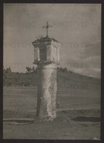 Vilnius vicinity. Chapel in Ponary. Photographer: Jan Bulhak 1876–1950. Village brick chapel, circa 1934. Place of origin: Vilnius Lithuania. Photograph nature photograph. Dimensions: height 15.7 cm, width 11.3 cm., A tall weathered stone column topped by a small rectangular niche structure with a metal cross stands in an open field; the column and niche show peeling and rough textures in shades of gray and off-white, with darker worn patches. The surrounding landscape features a gently sloping hill with a group of trees along its ridge and a grassy plain in front; the ground appears uneven with several shallow depressions. The sky above is cloudy with varying tones of gray. The overall image palette is monochromatic with tones of gray, off-white, and dark charcoal.