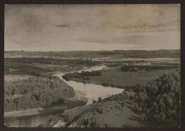 Ponary and Neris, Vilnius environs. Photographer: Jan Bulhak 1876–1950. Subject: Neris River, rivers, landscapes. Place: Vilnius, Lithuania. Date: ca. 1931. Medium: photography nature photography. Dimensions: 11.3 × 16.2 cm., A wide landscape photograph showing a river winding through a rural area with rolling hills and forested slopes; a group of trees with darker foliage occupies the foreground on both sides of the river, and lighter grassy fields lie beyond the river with scattered small groves and isolated trees; the distant horizon shows layered hills and a mix of open land and wooded areas under a broad sky with subtle cloud streaks. The visible colors are shades of black, gray, and muted brown.