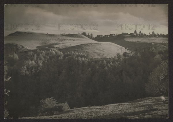 Ponarskie Mountains above the Wilja River Gorzuny, Jan Bulhak 1876–1950, photographer. Vilnius environment, Vilnius Lithuania. Nature photography; landscapes, hills. ca. 1934. Photography. Height 11.6 cm, width 16.6 cm. Purchase provenance., A landscape showing rolling hills and a wooded valley under a cloudy sky, with a group of houses along a ridgeline; visible colors include shades of black, gray, white, and muted sepia tones.
