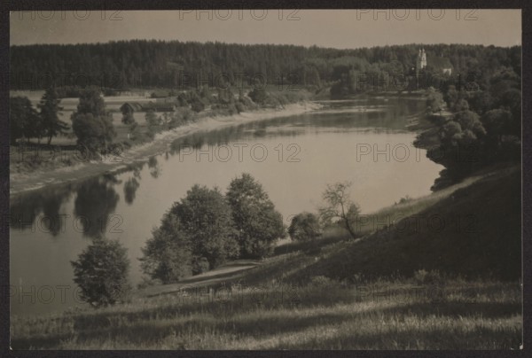 Trynopol; Bulhak, Jan 1876–1950, photographer; ca. 1937; photography nature photography; subject: landscapes — churches, rivers Neris, Verkiai; dimensions: 11.0 × 16.4 cm; Vilnius environs, Vilnius place of origin; provenance: purchase., A riverside landscape seen from a hillside with a calm, reflective body of water curving through the scene, grassy slopes in the foreground, a group of trees near the water’s edge including a few taller leafy trees and a more sparse tree with bare branches, a wooded shoreline on the opposite bank with a dense forest backdrop, several small structures partially visible among trees along the far bank, and a pale sky above; visible colors include shades of grayscale ranging from dark gray to light gray and muted sepia tones.