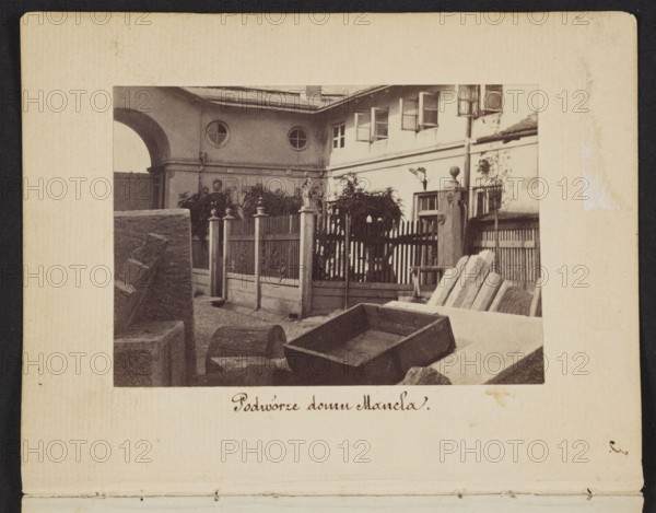 The Courtyard of the Mancel House View of the courtyard of the house and stonemason-sculptor workshop of Jan Józef Manzel, photographed by Karol Beyer 1818–1877; depicts Jan Józef Manzel 1806–1875. Date: 1861–1863. Medium: photographic print, nature photography/photography. Image: 5.5 cm high x 8 cm wide; support: 11.3 cm high x 8.8 cm wide. Location depicted: Warsaw, Chlodna Street, Masovian Voivodeship., A sepia-toned photograph shows a courtyard scene framed by a light-colored mat with a darker border. In the image, a building facade with a covered archway and round windows sits in the background, and a row of open rectangular windows is visible along the upper level. A wooden picket fence with decorative posts and finials runs across the middle ground, partially obscured by foliage and small trees. In the foreground there are several stone blocks and slabs, including a rectangular wooden or stone trough tilted on its side and a few angled slabs leaning against each other. Visible c...