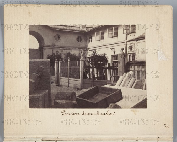 The Courtyard of the Mancel House View of the courtyard of the house and stonemason-sculptor workshop of Jan Józef Manzel, photograph by Karol Beyer 1818–1877; depicts Jan Józef Manzel 1806–1875 and his workshop; dates: 1861–1863; location: Chlodna, Warsaw Masovian Voivodeship; medium: photographic print, nature photography/photography; print dimensions: 5.5 cm x 8 cm; support dimensions: 11.3 cm x 8.8 cm., A sepia-toned photograph mounted on a beige card shows an outdoor scene with a courtyard enclosed by a wooden fence and gate. In the foreground there are several wooden and stone objects including a rectangular wooden box, a few planks or stone slabs leaning at an angle, and a rounded wooden barrel-like piece. Behind these objects a picket-style fence with vertical slats and decorative posts separates the courtyard from a building. The building has a pale facade with multiple windows, some open, and circular windows on an upper section. There is an archway on the left side of the bu...