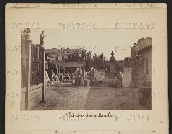 Courtyard of Mancel's House View of Jan Józef Manzel's stonemasonry and sculpting workshop, photograph by Karol Beyer 1818–1877; depicts Jan Józef Manzel 1806–1875, Léopold Méyet 1850–1912; subject: sculptors' workshop, tombstones; place: Chlodna, Warsaw, Masovian Voivodeship; date: 1861–1863; medium: photographic print nature photography; dimensions: print 5.7 × 9.2 cm; backing 11.3 × 8.8 cm., The image shows an outdoor courtyard scene with a cobblestone ground and a pathway leading between structures; there is a tall fence on the left with a statue mounted on a post and a group of rectangular stone blocks and slabs stacked and scattered toward the center and right, accompanied by a few upright pillars and carved stone elements; a few people are visible near the middle distance beside a cart and wooden beams, and there are buildings with pitched roofs and trees in the background; the visible colors include shades of brown, beige, tan, sepia, and muted gray.