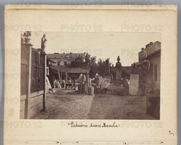 Courtyard of Mancel's House View of Jan Józef Manzel's stonemasonry and sculpting workshop, photograph by Karol Beyer 1818–1877; depicts Jan Józef Manzel 1806–1875. Subjects: sculptors' workshop, tombstones. Date: 1861–1863. Place: Chlodna, Warsaw Masovian Voivodeship. Medium/details: photographic print; nature photography; print size 5.7 × 9.2 cm; backing 11.3 × 8.8 cm., The image shows a courtyard scene bordered by a pale beige mat with a darker photograph centered within it; the photograph depicts an outdoor area with cobblestone ground, a gated wooden fence on the left topped by a small statue, and a collection of stone blocks and rectangular slabs arranged in the middle and right foreground. In the midground there is a group of people near a cart and more standing slabs, while the background includes a low building with a pitched roof, trees, and a few architectural elements such as pillars and a sculpture-like finial. Visible colors include shades of brown, beige, gray, black, an...