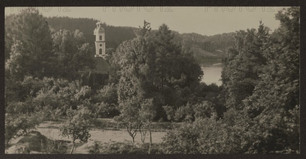 Trinopol and the Neris River, Bulhak, Jan 1876–1950, photographer; Trinopol Lithuania, churches; ca. 1937; photograph, nature photograph; 8.3 × 16.6 cm; Vilnius environs., A landscape dominated by a dense group of trees and bushes in shades of green, with a pale vertical tower partially visible among the foliage, a lighter-toned body of water peeking through the trees toward the right, a dark forested ridge in the background, and a foreground of grassy and sandy ground with a few small trees and shrubs; colors visible include various greens, pale off-white, light gray, sandy beige, and dark brown.