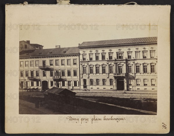 Houses at Bankowy Square Eastern frontage of Rymarska Street with Kruzy and Flatau's tenement houses, photograph by Karol Beyer 1818–1877, 1861–1863. Depicts Bankowy Square and Rymarska Street in Warsaw, showing Kruzy tenement house Rymarska 8 and Flatau's tenement house Rymarska 10. Photographic print nature photography, image: 6.2 × 9.5 cm; support: 11.3 × 8.8 cm., A sepia-toned photograph mounted on a cream-colored matte shows a street scene with a group of adjacent multi-story buildings featuring rows of windows and decorative architectural details; the building on the right has ornate window surrounds and a small balcony while the building on the left has simpler facades and balconies, and both have dark roofs. In front of the buildings there is a street with a few horse-drawn carriages and a darker wooden structure with a slanted roof in the foreground. Visible colors include cream, beige, brown, dark gray, and black.