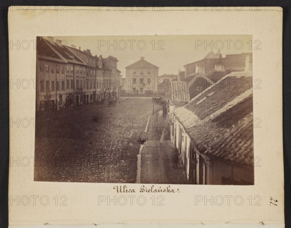 Bielanska Street View from Senatorska Street towards Dluga Street with the Arsenal building rebuilt into a prison Photographer/Author: Karol Beyer 1818–1877 Date: 1861–1863 Place: Warsaw; view includes Arsenal, Bielanska Street Subject/Notes: gutters; prison Medium: photography, photographic print Dimensions: print 6 cm h x 9.1 cm w; backing 11.3 cm h x 8.8 cm w, A sepia-toned photograph of a street scene framed by a wide cream border, showing a cobbled square with a group of multi-story buildings on the left and a row of lower buildings with tiled pitched roofs on the right; a larger square-fronted building stands at the far end of the square. The image shows a narrow sidewalk alongside the right-hand buildings, a few indistinct figures on the street and sidewalk, several shopfronts along the left-hand row, and a few street fixtures such as signs and lamp posts; visible colors include sepia-brown, cream, beige, and muted gray.