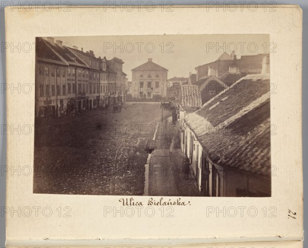 Warsaw. Bielanska Street View from Senatorska Street towards Dluga Street with the Arsenal building rebuilt into a prison, Beyer, Karol 1818–1877 author, view includes gutters, prison; dated 1861–1863; photography, nature photography; photographic print: 6 cm × 9.1 cm; backing: 11.3 cm × 8.8 cm., A sepia-toned photograph shows a street scene with a row of multi-story buildings on the left and a row of lower, tiled-roof buildings on the right, separated by a cobbled street and a narrow sidewalk. A large, boxy building stands at the end of the street in the background. The left buildings display multiple windows and shopfronts at ground level, while the right side features sloping roofs with visible tiles and small doorways. The street surface appears textured and uneven, and a few indistinct figures and a horse-drawn cart are visible along the roadway. The visible colors are shades of brown, beige, cream, and muted gray.