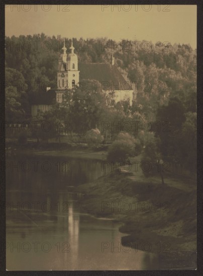 Vilnius environment. Trinopol, Bulhak, Jan 1876–1950 photographer, Vilnius Lithuania, landscapes, Trinopol Lithuania, rivers, churches, Neris Lithuania; river, ca. 1920, photography, nature photography, height 8.3 cm, width 16.6 cm, A sepia-toned scene shows a riverside landscape with a light-colored building featuring twin towers nestled among dense trees on a forested hillside, a group of trees and shrubs lining the riverbank, and the river in the foreground reflecting the lighter tones from the building and sky; visible colors include sepia/brown tones, darker brown and greenish hues in the trees, and lighter beige in the sky and building.