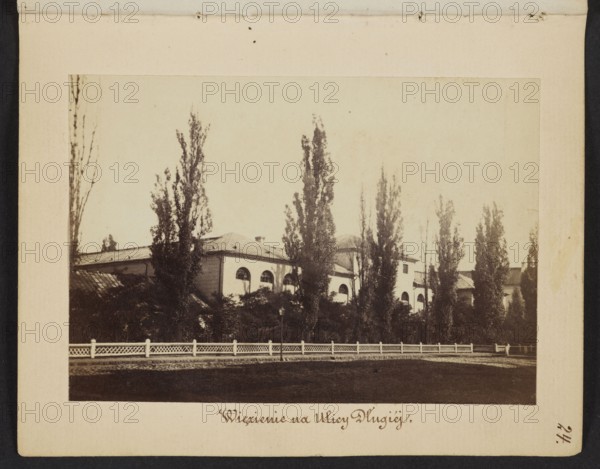 Prison on Dluga Street Arsenal Building Designated as a Prison, Beyer, Karol 1818–1877 author, Méyet, Leopold 1850–1912 photographer, Arsenal Warsaw, Dluga Warsaw; street, poplars, 1861–1863, Warsaw; photography, nature photography; photographic print: height 6.6 cm, width 9.4 cm; support: height 11.3 cm, width 8.8 cm, The image shows an outdoor scene with a pale beige background area and a darker rectangular photograph mounted on it; within the photograph there is a group of tall, narrow trees in dark brown tones standing in front of a light-colored building with arched windows and a pitched roof, partially obscured by darker foliage; a low white fence runs horizontally across the foreground above a darker strip of ground, and the overall color palette includes shades of beige, brown, white, and dark greenish-brown.