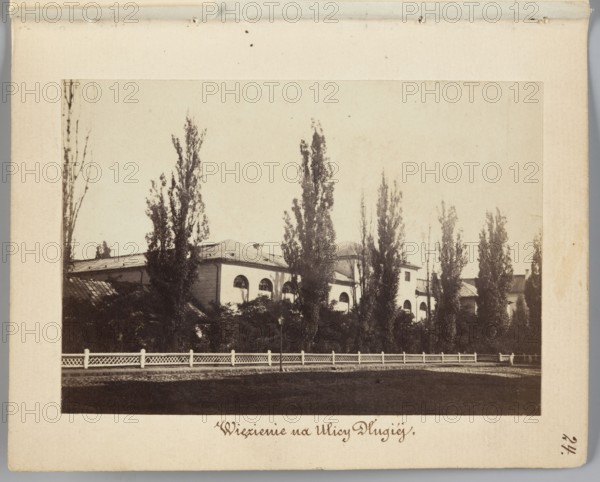 Warsaw. Prison on Dluga Street Arsenal Building Designated as a Prison, Beyer, Karol 1818–1877 author / photographer, prisons, Arsenal Warsaw, Warsaw Masovian Voivodeship, Dluga Warsaw; street, poplars, 1861–1863, Warsaw place of creation, photography, nature photography, photographic print: height 6.6 cm, width 9.4 cm, support: height 11.3 cm, width 8.8 cm, A sepia-toned scene showing a large light-colored building with arched windows partially obscured by a row of tall, narrow trees and dense dark foliage in front; a white decorative fence runs along the foreground with a dark paved area between the fence and the bottom edge of the image; the sky above is pale and mostly empty, and the overall colors visible are shades of brown, beige, cream, white, and dark greenish-brown.