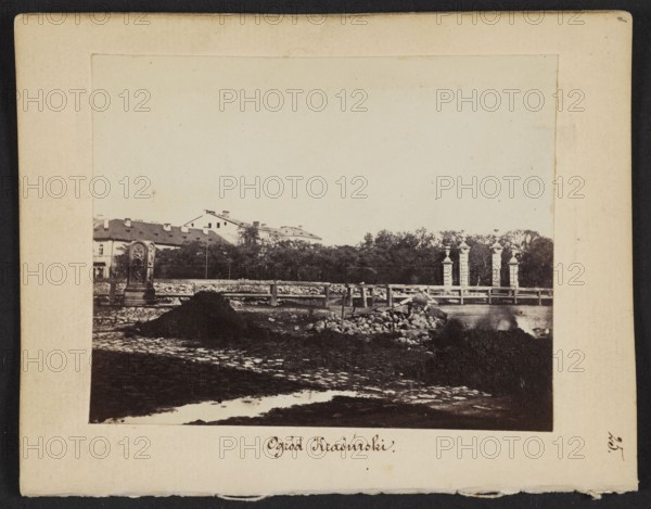 Krasinski Garden View of the gate to the Garden from Nalewki Street. On the left, buildings of Swietojerska Street, Beyer, Karol 1818–1877 photographer/author; subjects: garden gates, Krasinski Garden, cobblestones, Nalewki Street, Swietojerska Street; Marconi's waterworks, 1861–1863; photography, nature photography; photographic print: height 6.8 cm, width 8.6 cm; support: height 11.3 cm, width 8.8 cm., The image shows an outdoor scene framed by a pale border with a few darkened edges; within the scene there is a low wall or fence running horizontally, behind which a group of trees and a row of buildings with sloped roofs are visible. In front of the fence there is a paved or cobbled area with a few piles of dark soil or earth and a group of scattered rocks; a decorative stone monument or pillar stands to the left near the fence, and a set of taller stone columns or posts appears toward the right. The foreground contains patches of light reflecting on wet ground, and the overall color...