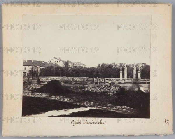 Krasinski Garden View of the gate to the Garden from Nalewki Street. On the left, buildings of Swietojerska Street, Beyer, Karol 1818–1877 photographer; garden gates; Krasinski Garden Warsaw; cobblestones; Nalewki Street Warsaw; Swietojerska Street Warsaw; waterworks; Marconi's waterworks Warsaw; 1861–1863; photography, nature photography; photographic print: height 6.8 cm, width 8.6 cm; support: height 11.3 cm, width 8.8 cm, A framed black-and-white photograph showing an outdoor scene with a muddy foreground and a few puddles, a dark mound and a pile of rocks near the center, a low stone or cobbled surface leading toward a wooden fence, a group of tall column-like structures with decorative tops beyond the fence, a line of trees behind them, a row of multi-roofed buildings partially visible above the trees to the left, and a carved stone monument or pillar near the left edge; visible colors include shades of black, white, gray, beige, and off-white.